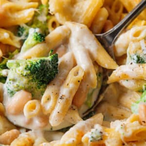 Close-up of baked penne pasta with broccoli, white beans, and a creamy alfredo casserole sauce, served with a metal spoon.