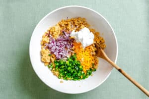 A white bowl with chopped red onion, cheddar cheese, jalapeno, cooked lentils, and a dollop of sour cream, with a wooden spoon on a light green surface.