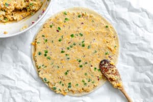 A round dough base topped with a mixture of cheese, chopped peppers, and onions sits on crumpled parchment paper, with a wooden spoon and a mixing bowl nearby.