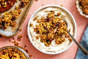 A bowl of yogurt topped with granola and seeds sits on a pink surface, with a spoon in the bowl and more granola scattered nearby.