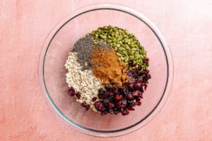 A glass bowl containing oats, dried cranberries, pumpkin seeds, chia seeds, and ground cinnamon on a pink surface.