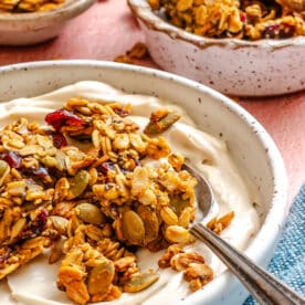 A bowl of yogurt topped with granola and seeds, with a spoon resting inside. Two additional bowls of granola are in the background.