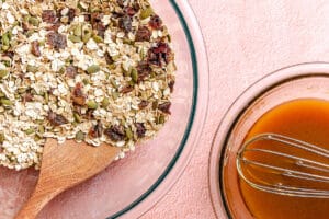 A glass bowl of dry granola ingredients with a wooden spoon next to a bowl of orange liquid mixture with a whisk, on a pink surface.
