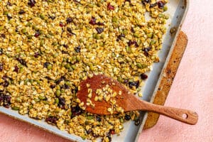 A baking tray filled with homemade granola containing oats, seeds, and dried fruit, with a wooden spoon resting on top.