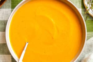 A bowl of smooth orange soup with a spoon, placed on a green and white checkered tablecloth.