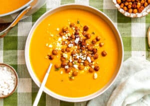 A bowl of smooth orange soup topped with roasted chickpeas and crumbled cheese, with a spoon resting inside, on a plaid tablecloth.