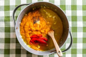 A pot containing chopped onions, pureed pumpkin, sliced red bell pepper, spices, and broth sits on a green and white checkered tablecloth with a wooden spoon inside.