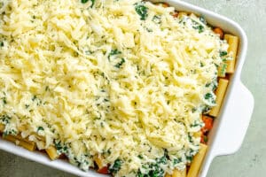 A white baking dish filled with uncooked manicotti pasta, spinach, tomato sauce, and topped with a generous layer of shredded cheese.