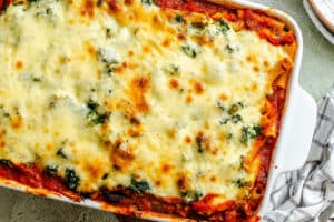A close-up of a baked pasta casserole with melted cheese, tomato sauce, and visible herbs in a white rectangular dish.