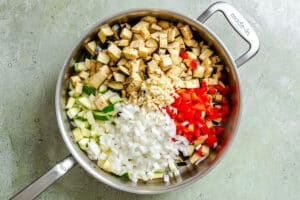 A stainless steel pan contains chopped eggplant, zucchini, onion, red bell pepper, and minced garlic, arranged in separate sections on a light gray surface.