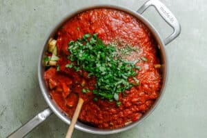 A large pan filled with tomato sauce, chopped fresh herbs, and vegetables, being stirred with a wooden spoon.