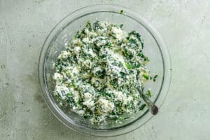 A glass bowl containing a mixture of ricotta cheese, chopped spinach, and herbs with a spoon resting inside.