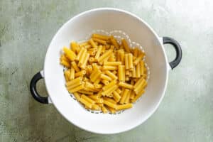 Uncooked ziti pasta in a white colander with black handles, placed on a light green textured surface.
