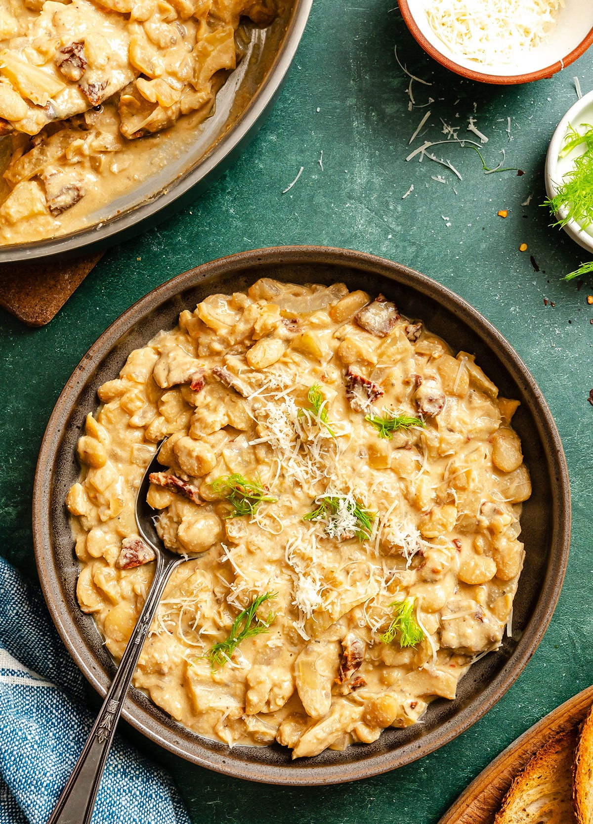 A bowl of creamy pasta with pieces of meat, topped with grated cheese and herbs, sits on a green surface next to a pan of the same dish and some bread.