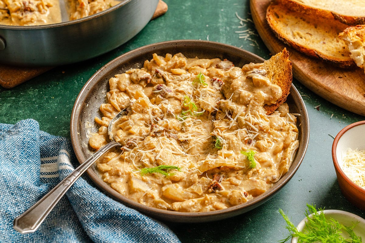 A bowl of creamy braised butter beans with shredded cheese, sun-dried tomatoes, and a slice of toasted bread, served on a green surface next to a blue napkin and extra grated cheese.