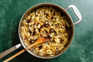 A skillet on a green countertop with sautéed chopped fennel, onions, and sun-dried tomatoes being stirred with a wooden spoon.