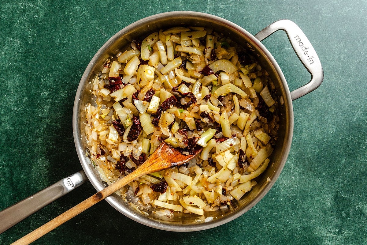 A skillet on a green countertop with sautéed chopped fennel, onions, and sun-dried tomatoes being stirred with a wooden spoon.