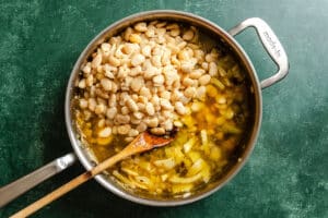 A saucepan filled with white beans, broth, and chopped vegetables sits on a green surface with a wooden spoon inside.
