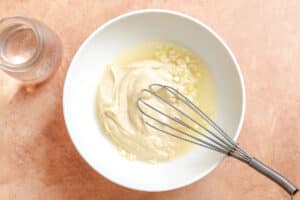 A white bowl contains tahini, lemon juice, chopped garlic, and a whisk on a peach-colored surface, with a glass jar nearby.