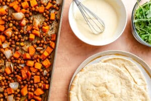 Sheet pan of roasted chickpeas, sweet potatoes, and onions next to a bowl of white sauce with a whisk, a plate with pita bread, and a bowl of fresh arugula.