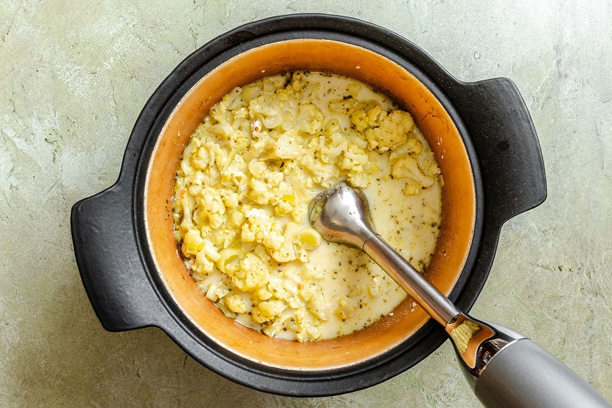 A pot of cooked cauliflower in a creamy liquid sits on a light surface with an immersion blender partially submerged in the mixture, ready for blending.