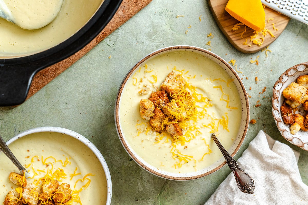 Two bowls of creamy cauliflower soup topped with croutons and shredded cheese sit on a green surface, with a pot of soup and a cutting board with cheese nearby.