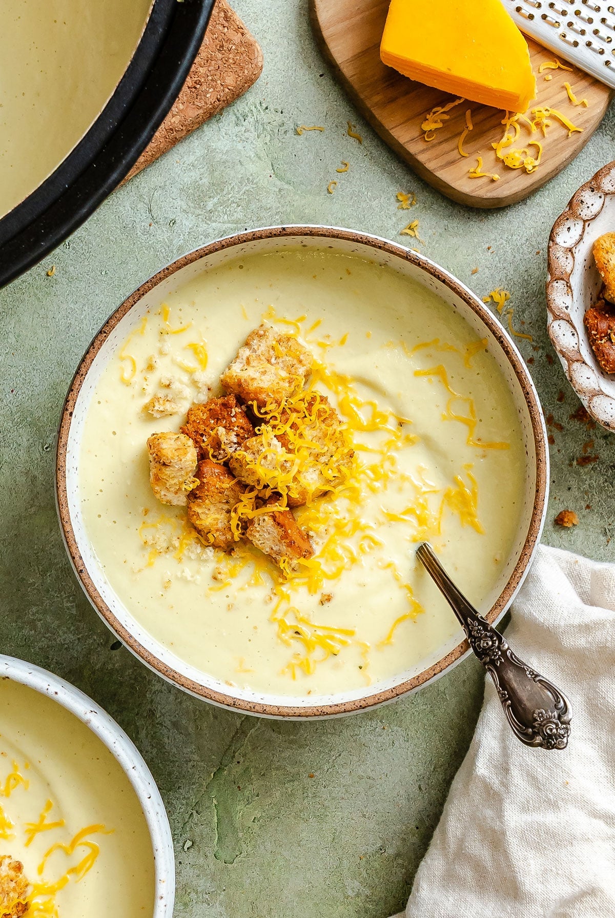 Bowl of creamy cauliflower soup topped with shredded cheese and croutons, with a spoon inside. Nearby are more croutons and a block of cheese on a cutting board.
