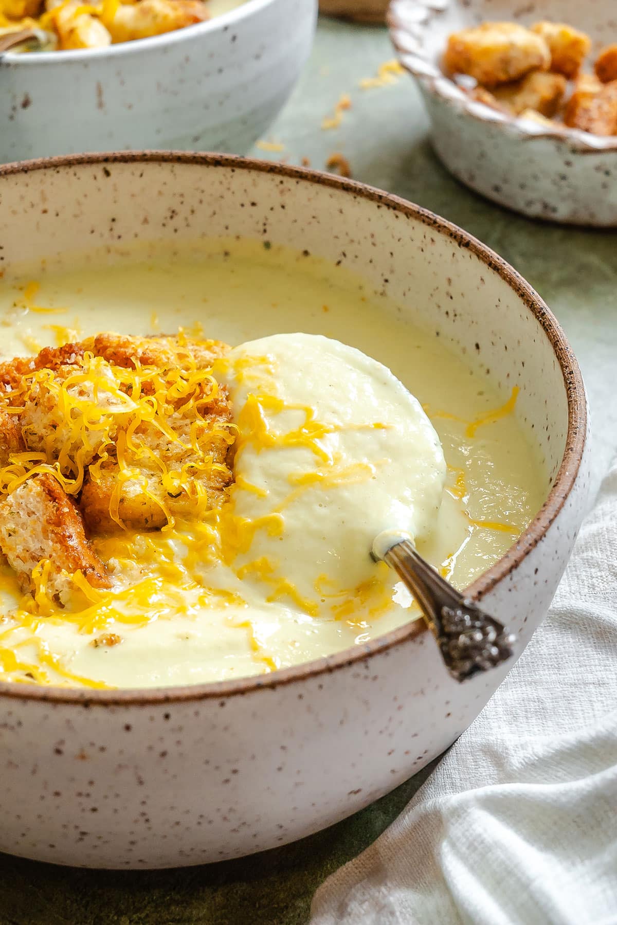 A bowl of creamy cauliflower soup topped with shredded cheese and croutons, with a spoon resting inside.