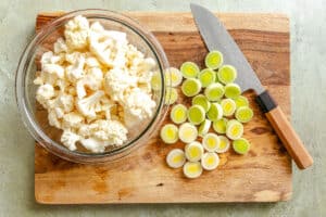 A glass bowl of cauliflower florets and sliced leeks on a wooden cutting board with a knife placed beside them.