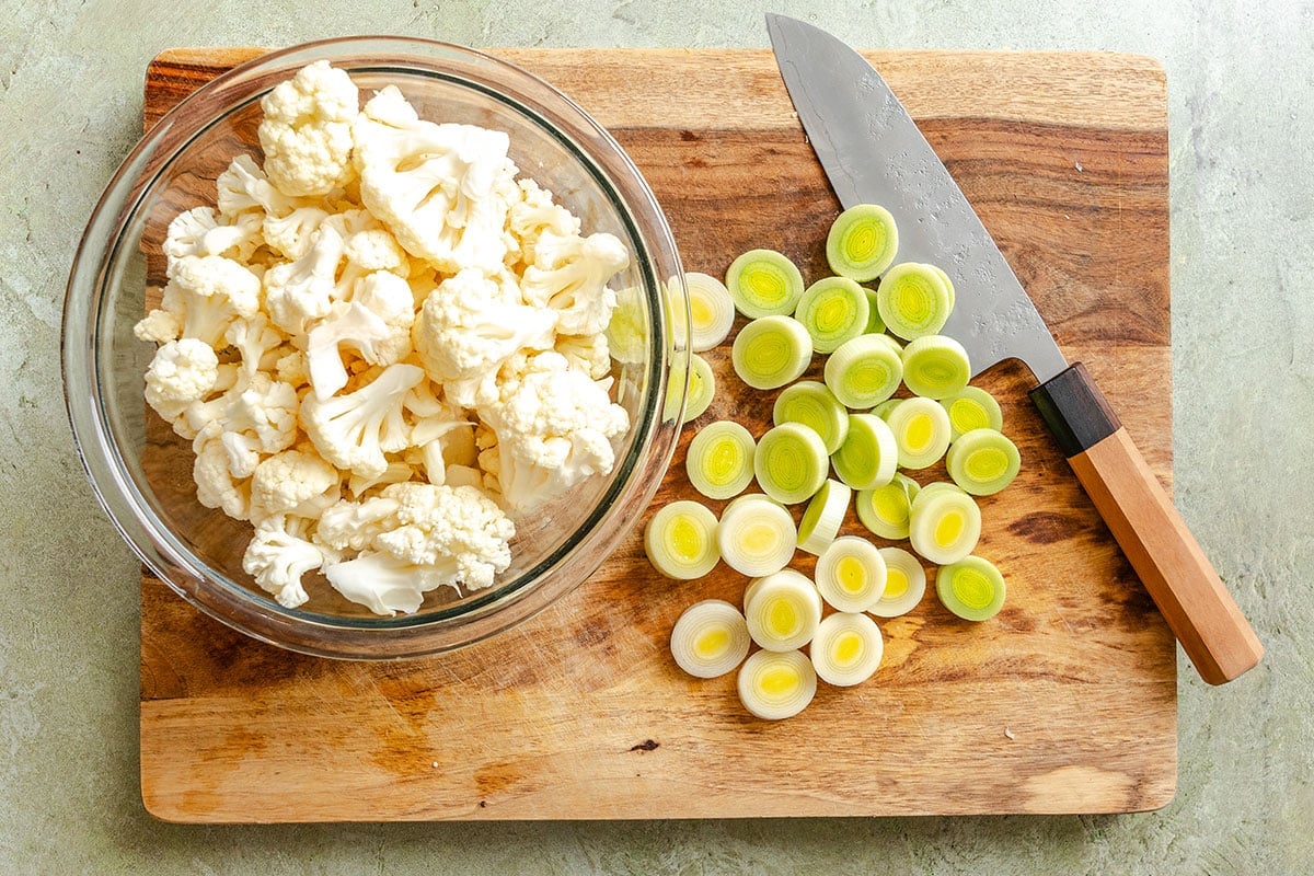 A glass bowl of cauliflower florets and sliced leeks on a wooden cutting board with a knife placed beside them.