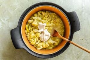 A black pot containing cooked cauliflower, yellow sauce, light liquid, and a dollop of cream with a wooden spoon resting inside.