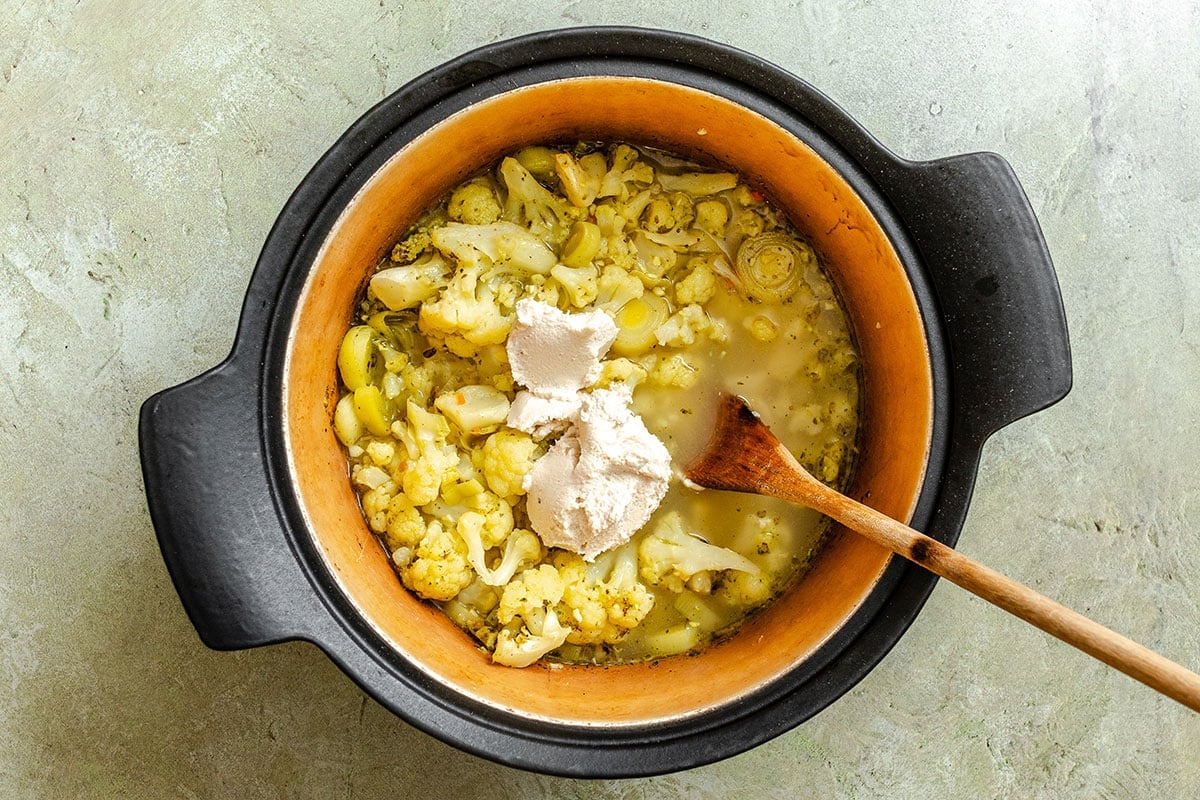 A black pot containing cooked cauliflower, yellow sauce, light liquid, and a dollop of cream with a wooden spoon resting inside.