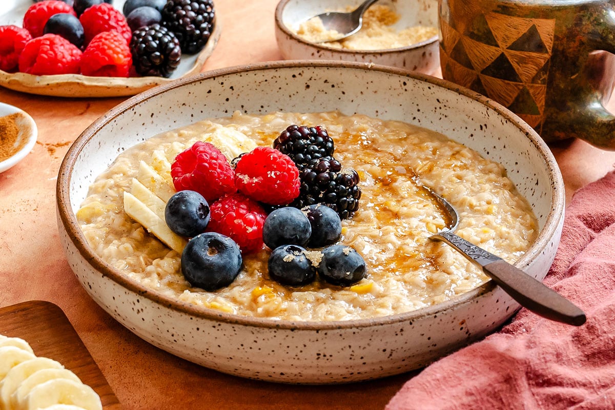 A bowl of chickpea oatmeal topped with banana slices, blueberries, raspberries, and blackberries, with a spoon and a drizzle of syrup. Plates of berries and a mug are in the background.