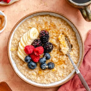 A bowl of oatmeal topped with sliced bananas, raspberries, blackberries, and blueberries, surrounded by ingredients like fresh fruit, brown sugar, cinnamon, and a cup of coffee.
