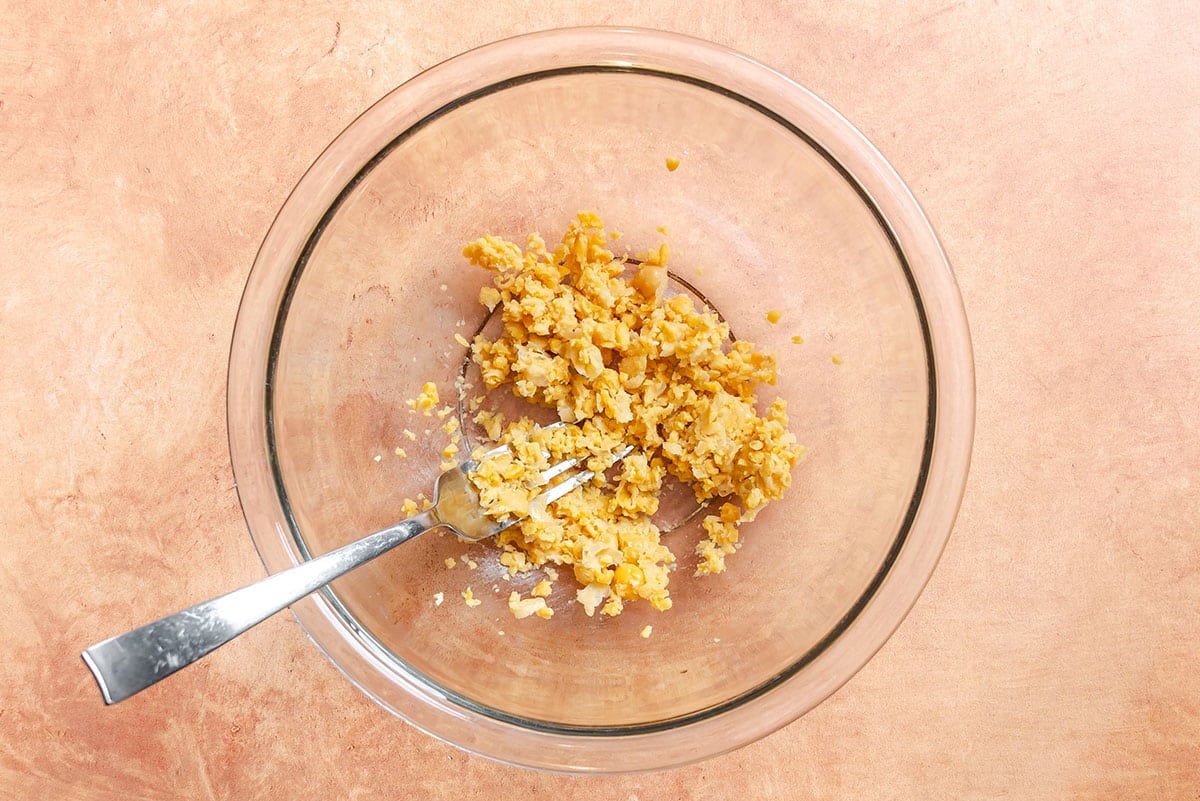 A glass bowl containing mashed chickpeas with a fork, placed on a light brown surface.