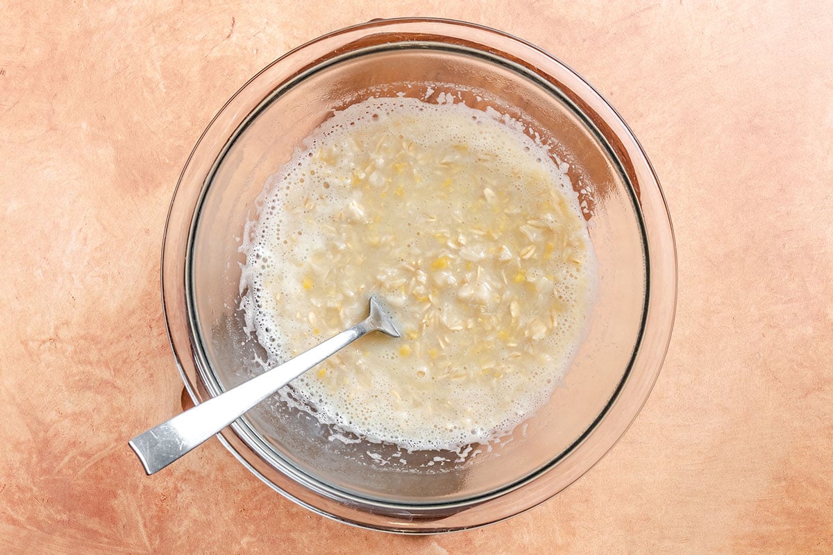 A glass bowl with mashed bananas and a silver fork sits on a beige countertop.
