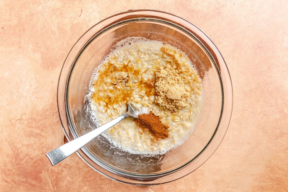 A glass bowl with oats, milk, brown sugar, cinnamon, and vanilla being mixed with a spoon on a light brown surface.