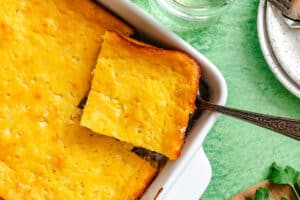 Golden-brown casserole with a square piece being served from a white baking dish, on a green surface with plates and parsley nearby.