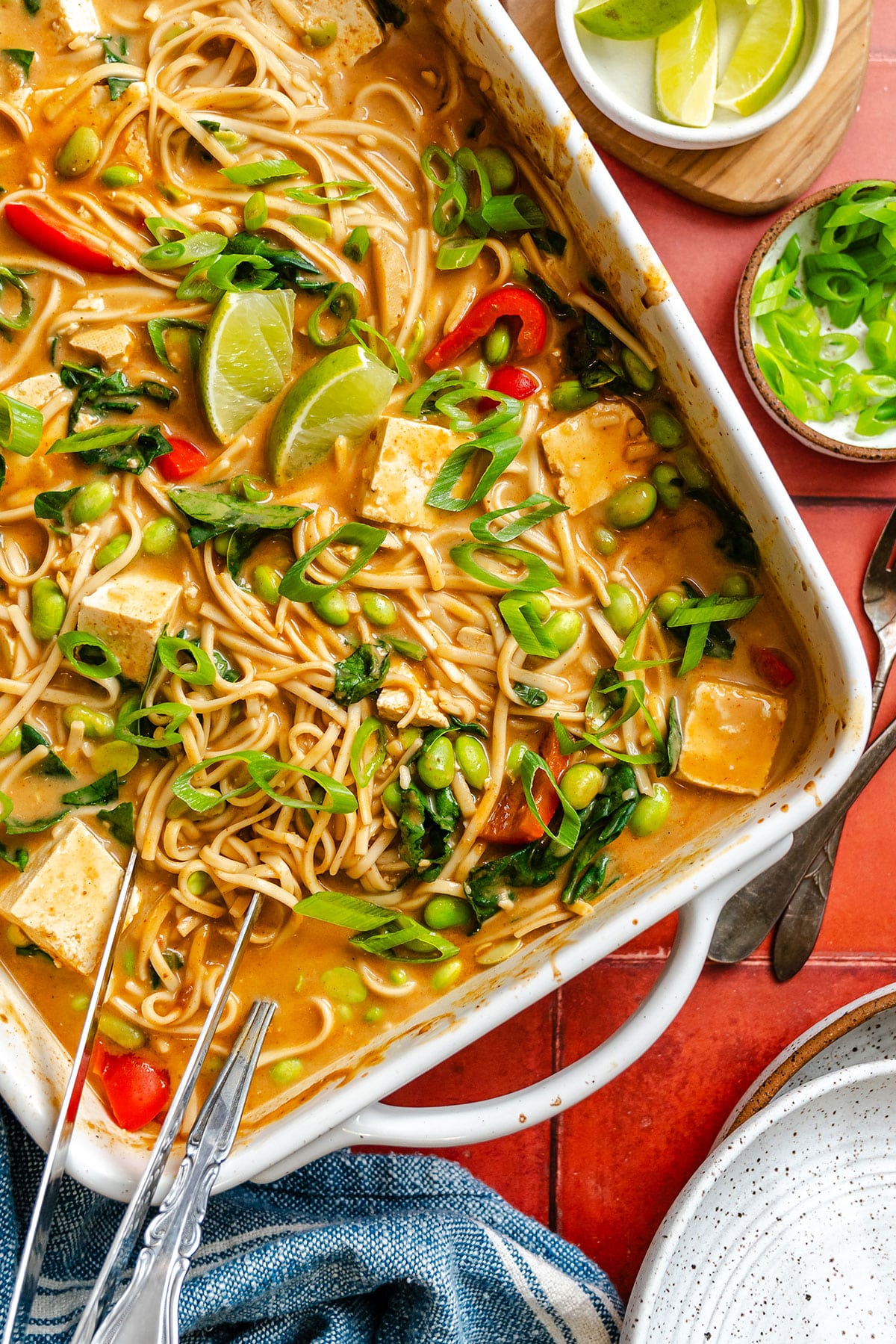 A white casserole dish filled with noodle soup containing tofu, edamame, greens, and red peppers, garnished with lime wedges and chopped scallions. Lime slices and scallions on the side.