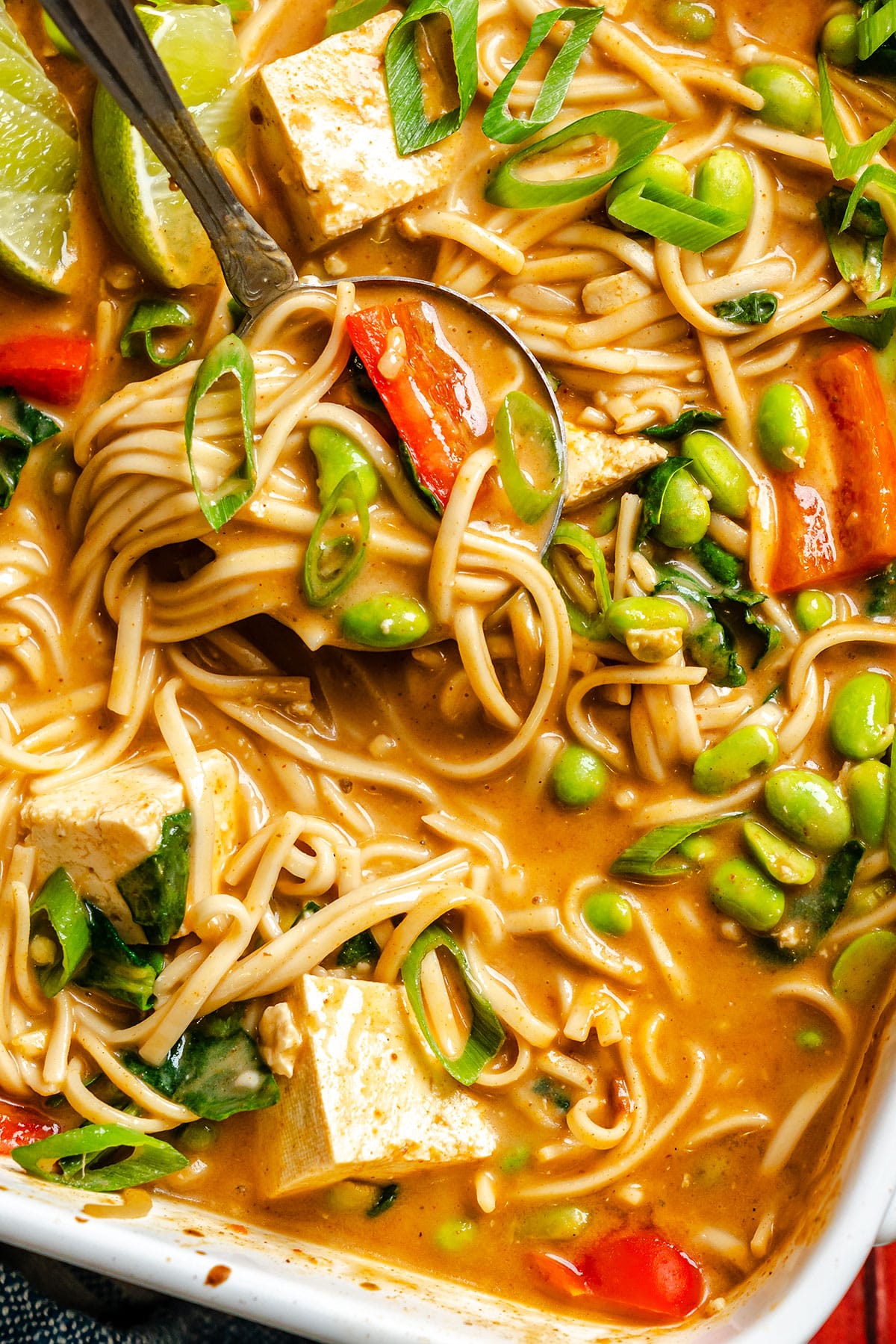 A close-up of a bowl of noodle soup with tofu, edamame, red peppers, chopped scallions, lime wedges, and a creamy broth, with a spoon and fork in the dish.