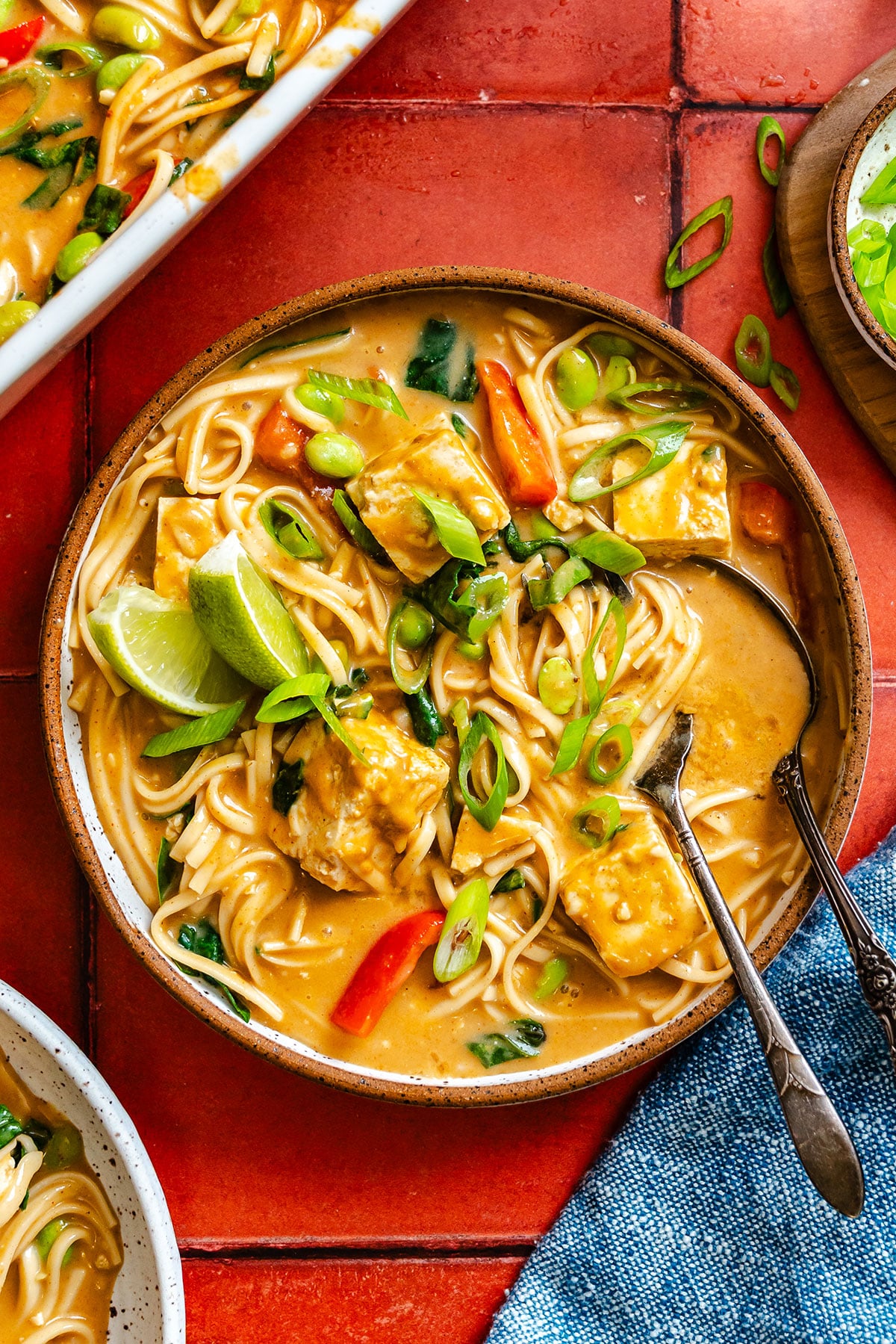 A bowl of noodle soup with vegetables, tofu, lime wedges, and chopped green onions, served with two spoons on a red tiled surface.