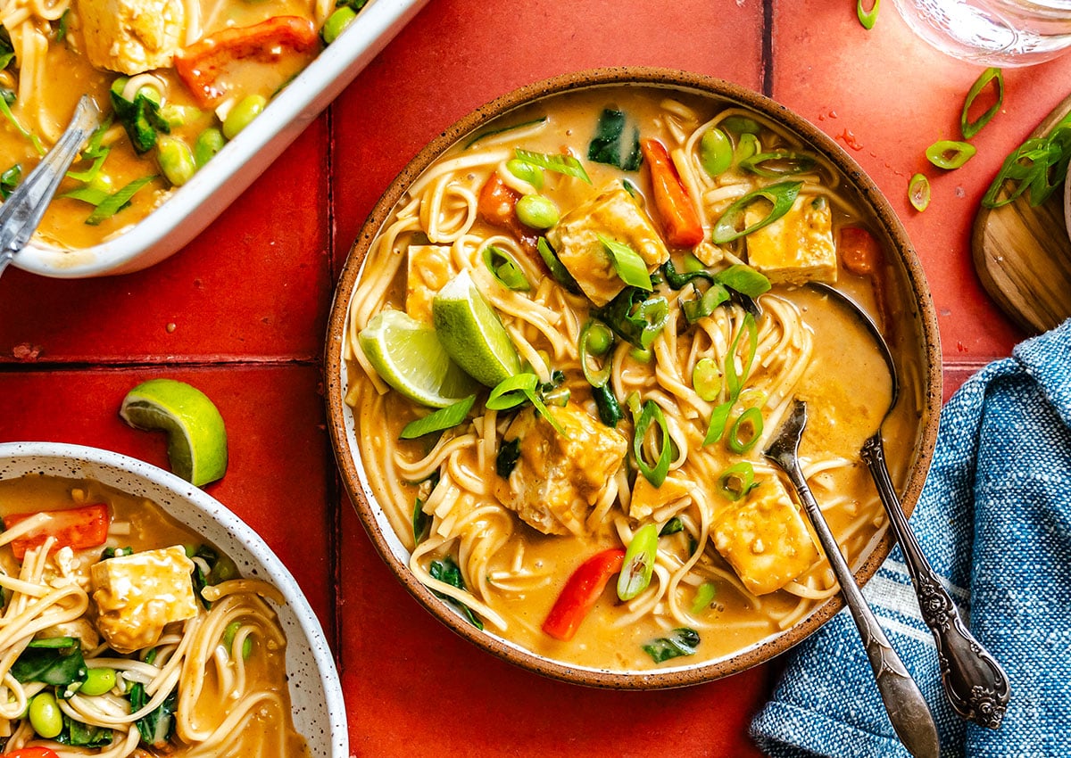 A bowl of noodle soup with tofu, red peppers, greens, and scallions, garnished with lime wedges, placed on a red tiled surface with a blue napkin and additional dishware nearby.