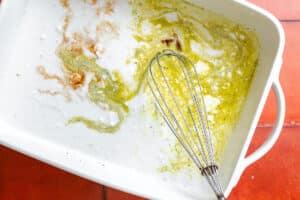 A white baking dish with remnants of batter and a metal whisk, sitting on a red-tiled surface.