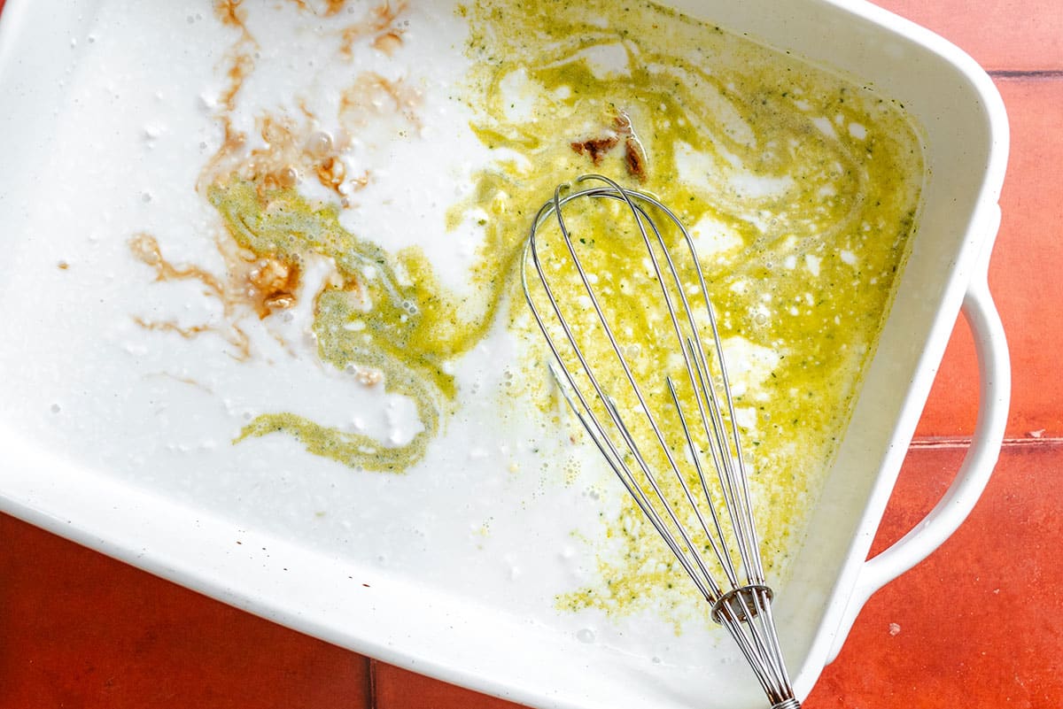 A white baking dish with remnants of batter and a metal whisk, sitting on a red-tiled surface.