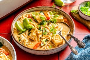 A bowl of noodle soup with tofu cubes, sliced green onions, red bell peppers, and lime wedges, served on a red tile surface with a fork in the bowl.