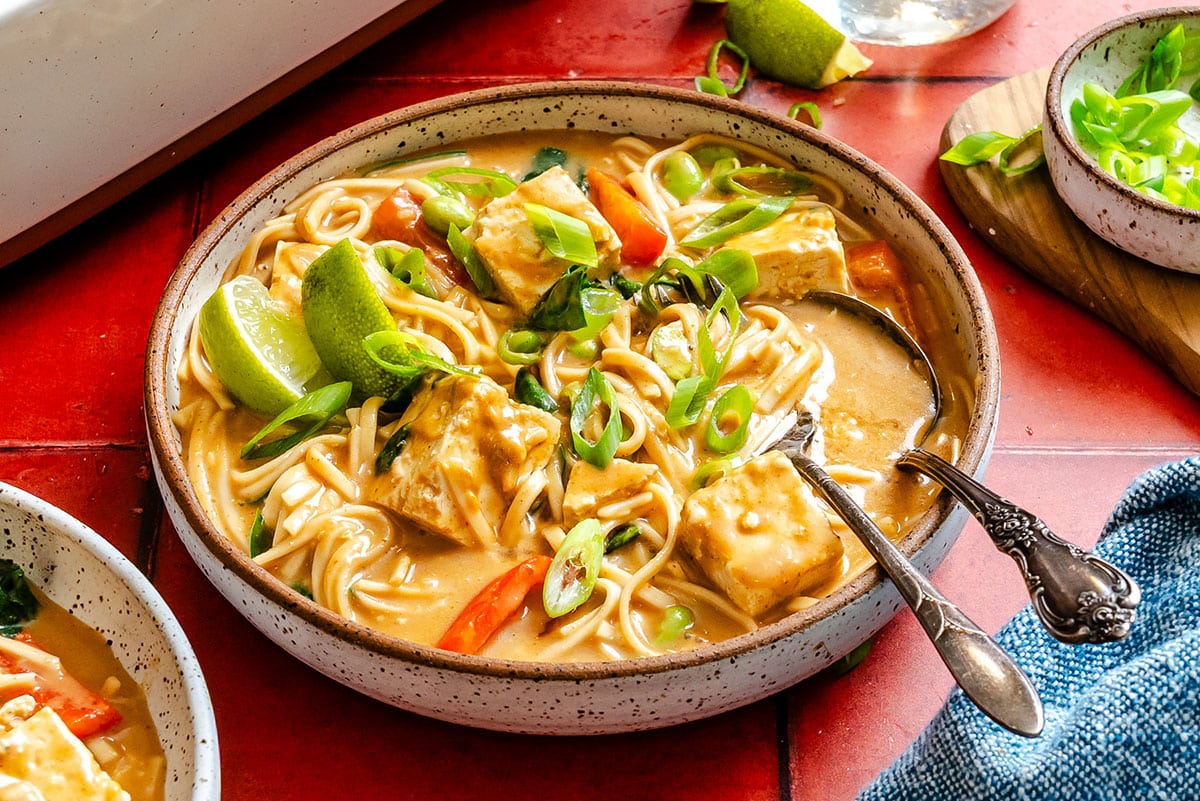 A bowl of noodle soup with tofu cubes, sliced green onions, red bell peppers, and lime wedges, served on a red tile surface with a fork in the bowl.