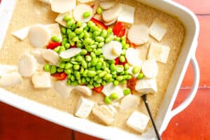 A white dish filled with tofu cubes, sliced water chestnuts, edamame, red bell pepper pieces, and a light-colored sauce, with a spoon partially visible.