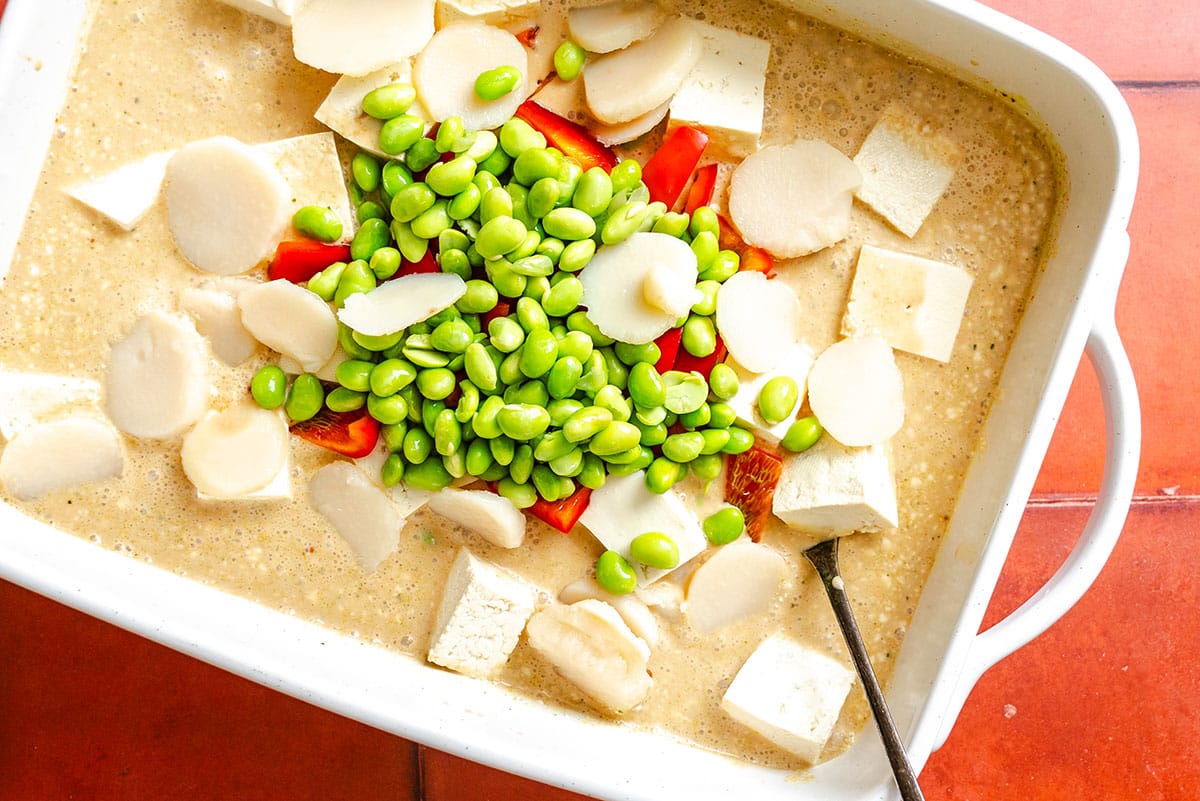 A white dish filled with tofu cubes, sliced water chestnuts, edamame, red bell pepper pieces, and a light-colored sauce, with a spoon partially visible.