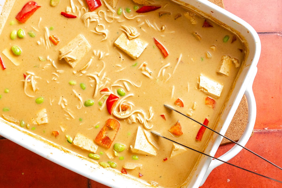 A casserole dish filled with soup containing tofu cubes, red bell pepper pieces, green edamame, and noodles in a creamy broth.