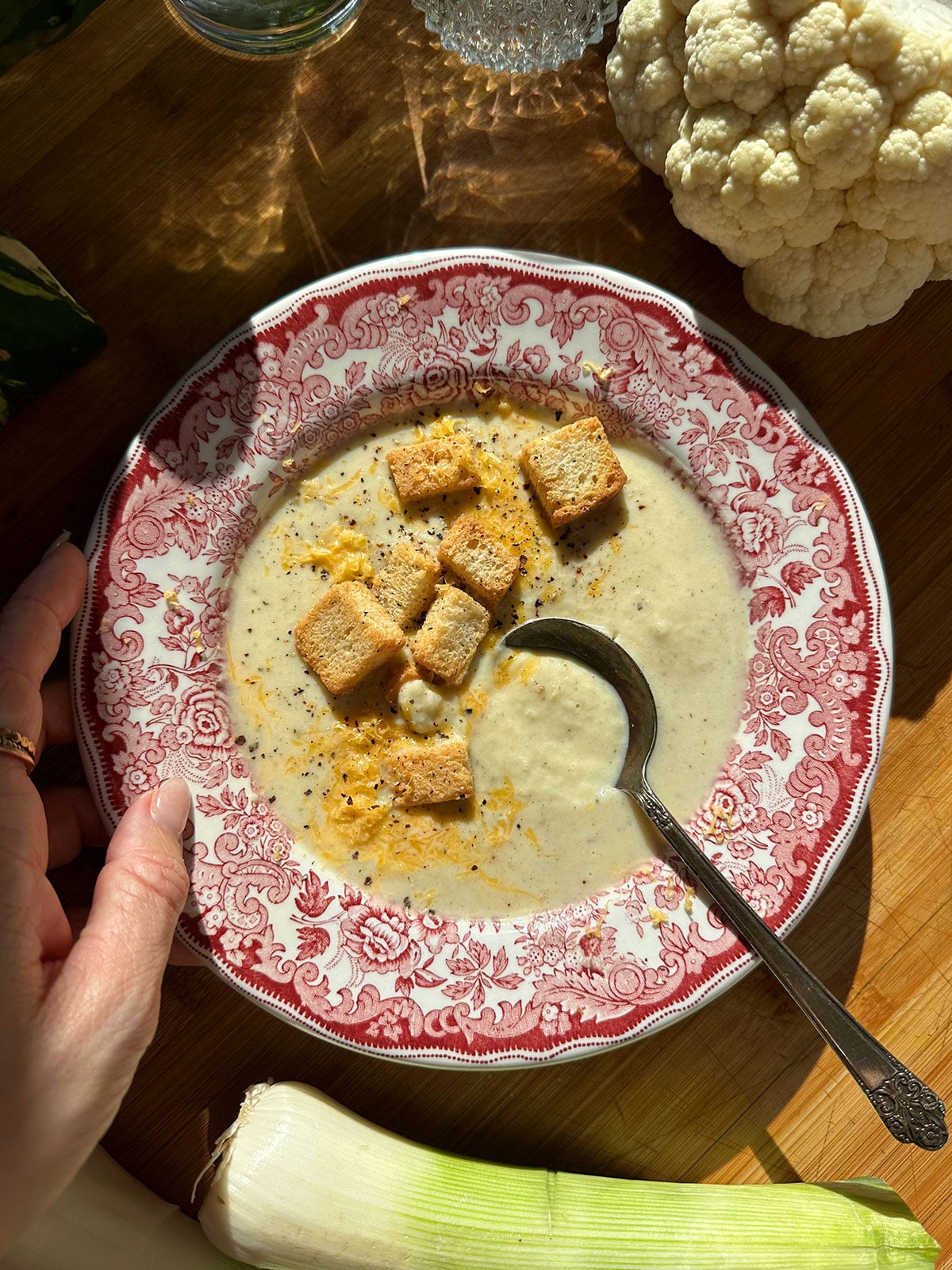 A bowl of creamy soup topped with croutons and pepper sits on a patterned plate with a spoon. Cauliflower and a leek are nearby on a wooden surface. A hand holds the plate.
